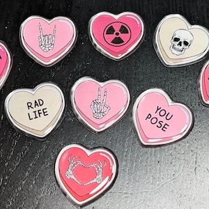 May include: Eight pink and white heart-shaped candy conversation hearts with various skeleton and skull designs and phrases including "Skeleton Crew", "Rad Life", "We Expose", "You Pose", and "Peace" signs.