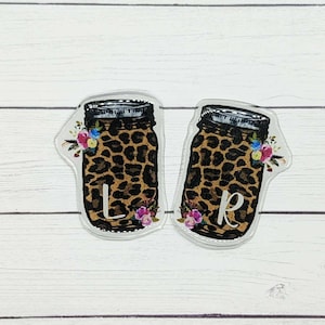 May include: Two clear acrylic mason jar ornaments with a leopard print design and floral accents. The jars are labeled "L" and "R".