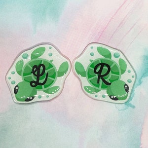 May include: Two green turtle resin charms with white outlines and black eyes. The turtles have a white shell with a black letter on each. The left turtle has the letter "L" and the right turtle has the letter "R".