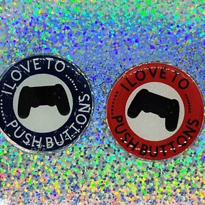 May include: Two round buttons with the text "I love to push buttons" and a black and white video game controller graphic. One button is blue and the other is red.