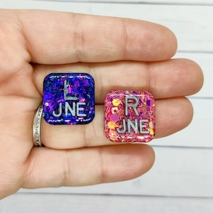 May include: Two square resin pieces with glitter. One is dark blue with the letter "L" and the word "JUNE" in silver. The other is pink with the letter "R" and the word "JUNE" in silver. The pieces are held in a hand.