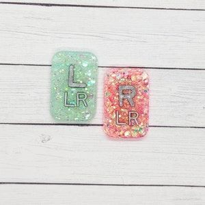 May include: Two small, rectangular, glitter-filled resin charms with the letters "L" and "R" and the abbreviation "LR" printed on them. The charm on the left is green and the charm on the right is pink.