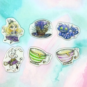 May include: A collection of six clear acrylic charms featuring illustrations from Alice in Wonderland. The charms include Alice, the Mad Hatter's hat, the Cheshire Cat, the White Rabbit, and two teacups with striped designs.