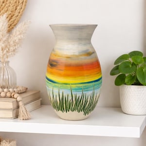 May include: A tall, ceramic vase with a painted sunset design. The vase features a gradient of colors, including orange, yellow, and blue, with green grass at the base. The vase is displayed on a white shelf.