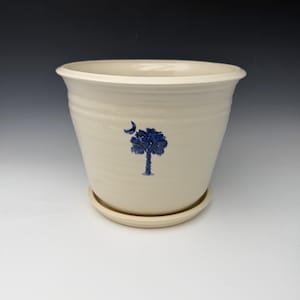 May include: Cream-colored ceramic planter with a matching saucer. The planter features a blue tree and crescent moon design. The pot has a wide opening and a slightly textured surface. A decorative item for indoor or outdoor use.