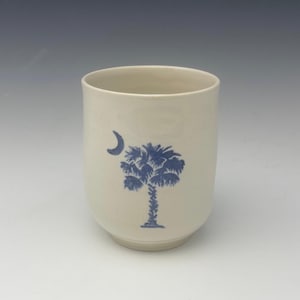 Puede incluir: Una taza de cerámica blanca con un diseño de palmera azul y media luna.