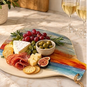 May include: A teardrop-shaped serving board with a painted abstract design in blue, orange, and white. It holds a spread of brie cheese, salami slices, crackers, green olives in a small bowl, red grapes, fresh figs, and rosemary sprigs.