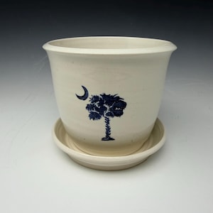 Może przedstawiać: Kremowy ceramiczny doniczka z pasującym spodkiem. Doniczka ma ciemnoniebieski wzór półksiężyca i palmy. Doniczka ma szeroką krawędź i lekko zwężający się kształt. Spodek jest okrągły i pasuje pod spód.