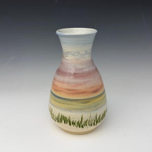 May include: A hand-painted ceramic vase with a landscape design. The vase features a gradient of colors, including blue, pink, orange, and yellow, with green grass painted at the base. The vase is approximately 8 inches tall.