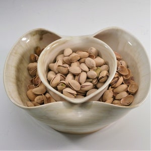 May include: A white ceramic serving dish with three sections, filled with pistachios. The dish is shaped like a heart and has a rustic, handmade look.