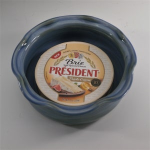 May include: A blue ceramic bowl with a wavy rim, containing a round package of President Brie Triple Creme cheese. The package is yellow with red and white lettering and features a picture of a wedge of brie cheese.