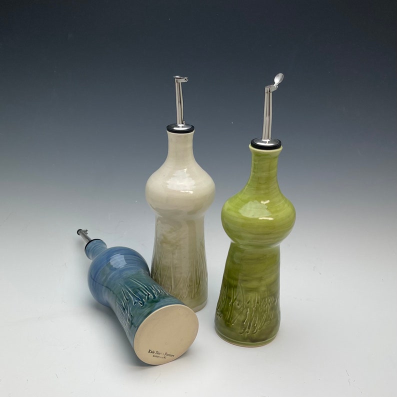 Op de afbeelding: Drie keramische olie- en azijnflessen met een textuurontwerp. De flessen zijn in tinten groen, blauw en wit. Elke fles heeft een giettuit van roestvrij staal.