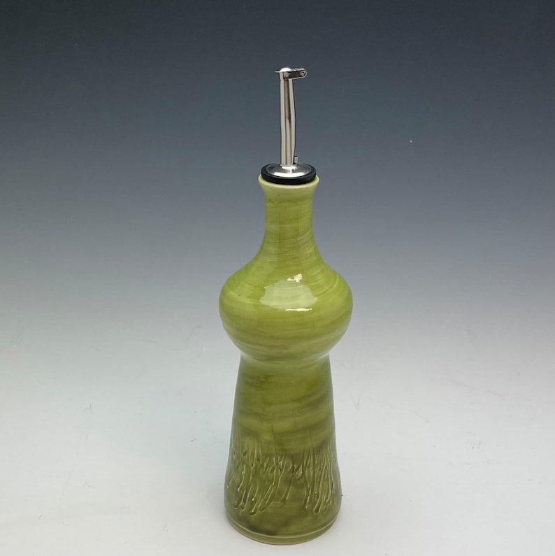 Op de afbeelding: Een groene keramische fles voor olie en azijn met een roestvrijstalen giettuit. De fles heeft een getextureerd, swirl-ontwerp.
