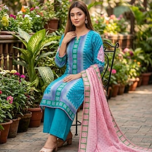 May include: A turquoise long-sleeved tunic with a central green and white geometric pattern, paired with matching turquoise wide-leg trousers. A light pink shawl with a green and white border and a subtle floral print drapes over the lap.