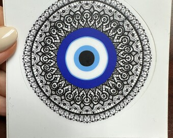 May include: A black and white mandala with a blue evil eye in the center. The evil eye is surrounded by a white circle and a blue circle. The mandala is on a white background.