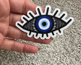 May include: A black and white sticker with a blue and white evil eye design. The eye is surrounded by a black and white pattern.
