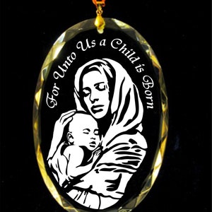 Puede incluir: Adorno ovalado de cristal con borde dorado y fondo negro. El adorno presenta un gráfico blanco de una madre sosteniendo a un bebé, con el texto "For Unto Us a Child is Born."