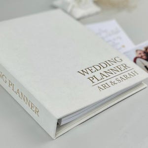 May include: A white wedding planner book with gold foil lettering on the front cover and spine. The cover text reads WEDDING PLANNER ARI & SARAH. The book has a hard cover and contains white paper pages visible along the side edge of the planner.
