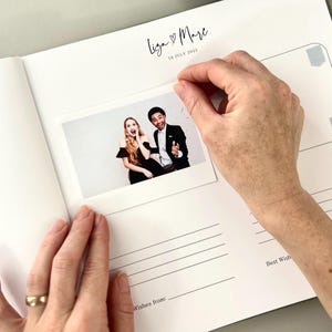 May include: A white wedding guest book with the names "Liza & Marc" and the date "19 July 2025" printed in black. A photo is being placed inside the book. The photo shows a smiling couple. The book has lined pages for messages.