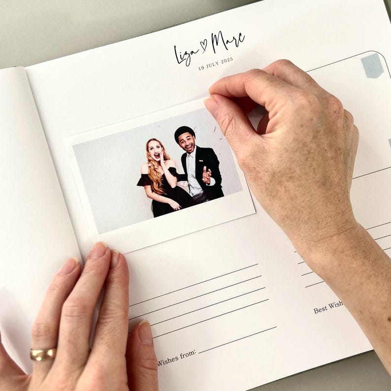 May include: A white wedding guest book with the names "Liza & Marc" and the date "19 July 2025" printed in black. A photo is being placed inside the book. The photo shows a smiling couple. The book has lined pages for messages.