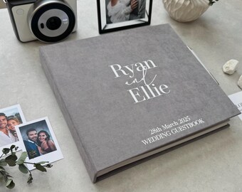 May include: A square, gray fabric-covered wedding guestbook with white text that reads Ryan and Ellie, 28th March 2025, WEDDING GUESTBOOK. The book is positioned on a surface next to an instant camera, a framed photograph, and small polaroid prints.