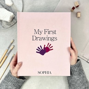 Op de afbeelding: Een lichtroze hardcover boek met de titel "My First Drawings" en een paars en magenta handafdrukontwerp. De naam "SOPHIA" staat onderaan gedrukt. Het boek wordt vastgehouden door een persoon met een grijze trui. Penselen en stempels staan op de achtergrond.