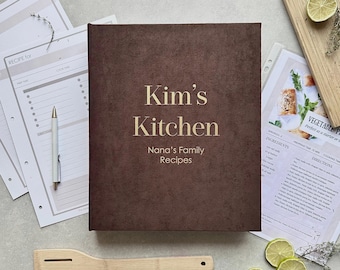 May include: A dark brown, textured recipe binder with the gold-colored text "Kim's Kitchen" and "Nana's Family Recipes" printed on the front cover. The binder is surrounded by recipe cards, a white pen, a wooden spatula, and lime slices.