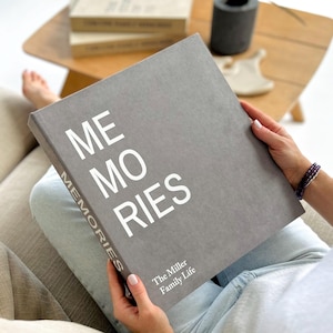 May include: A gray photo album with the word "MEMORIES" in large white letters. The spine reads "MEMORIES" vertically. The text "The Miller Family Life" is printed at the bottom. The album is held in a person's hands.