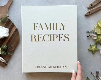 May include: A rectangular book with a light cream-colored fabric cover featuring the dark brown text FAMILY RECIPES centered in the upper half and the name LEBLANC-MCKEIGHAN printed in smaller dark brown text near the bottom edge.