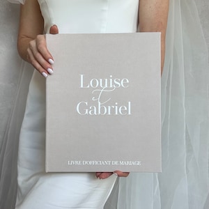 Puede incluir: Un libro de visitas de boda de lino beige con los nombres "Louise & Gabriel" impresos en blanco en letra cursiva. El texto "Livre d'officiant de mariage" está impreso en blanco en la parte inferior del libro.