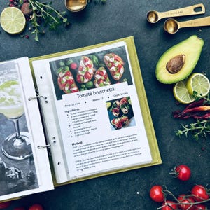 An opened binder showing transparent sleeves of recipe sheets printed from the internet. There are printed recipes as an example. A5 size 2 rings binder. The binder is on black table surrounded cherry potatoes, avocadoes, rose marines and lime.