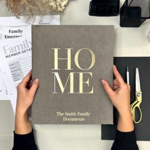 May include: A grey family documents binder with the word "HOME" in gold lettering. The text "The Smith Family Documents" is below the word "HOME". The binder is held by two hands. Other items on the desk include paperwork, scissors, and a pen.
