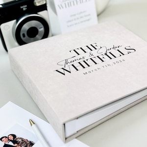 May include: A light-colored wedding photo album with the names "Thomas & Jordan Whitfills" and the date "March 7th, 2026" printed in black. A black and white instant camera and a pen are visible.