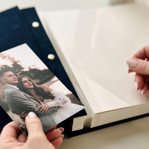 Puede incluir: Un álbum de fotos de terciopelo azul marino oscuro con cierres metálicos dorados. El álbum cuenta con páginas gruesas de color crema. Una fotografía rectangular que muestra a una pareja con ropa de boda está siendo colocada en el álbum.