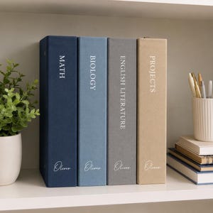 May include: Four vertical binders in navy, light blue, gray, and tan with white text labels reading MATH, BIOLOGY, ENGLISH LITERATURE, and PROJECTS. Each binder features the name Oliver in white script at the bottom of the spine.