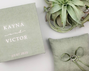 May include: A green ring pillow with a satin ribbon and two silver rings. The pillow is made of a textured fabric. A green box with the text "KAYNA and VICTOR 03 07 2020" is also in the image.