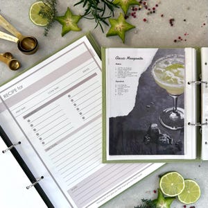 An opened recipe book page with a pink header that reads 'RECIPE for'. The recipe book page has sections for prep time, cook time, serves, ingredients, directions, and notes. And printed cocktail recipe from the internet as an example.