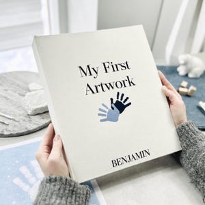 May include: A hardback book with a cream cover, titled "My First Artwork" with the name "BENJAMIN" below. The cover has a handprint design in black and light blue. The book is held by a person.