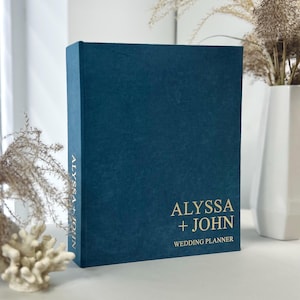 May include: A teal wedding planner with gold lettering. The cover reads "ALYSSA + JOHN WEDDING PLANNER." The spine has the name "ALYSSA + JOHN" printed vertically. The planner is standing upright.