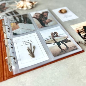 Könnte beinhalten: Ein braunes Lederfotoalbum mit durchsichtigen Plastikhüllen zum Aufbewahren von Fotos. Das Album ist geöffnet und zeigt Fotos eines Paares am Strand, eines Paares auf einem Surfbrett und ein Zitat, das lautet: "Das Leben ist eine Sammlung von Momenten".