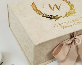 May include: A beige velvet gift box with a gold foil design featuring a wreath and the letter "W". The box is tied with a beige satin ribbon and has the text "CHINA & BRETT February 23, 2019" printed on the top.