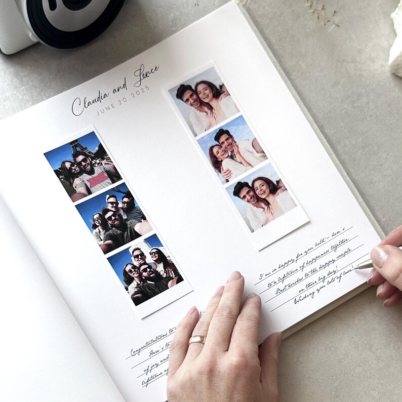May include: A white wedding photo album with the names "Claudia and Jevce" and the date "JUNE 20.2025" printed on the top. The album contains photo strips and handwritten notes. A hand with a gold ring is writing on the page.