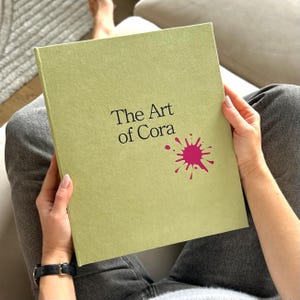 May include: A light green hardcover book titled "The Art of Cora" with a pink paint splatter design. The book is held open by a person sitting on a couch. The person is wearing a black watch and gray pants.