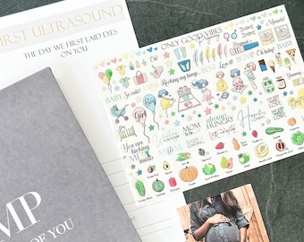Op de afbeelding: Een witte kaart met kleurrijke stickers met babygerelateerde illustraties en tekst zoals "Baby So Cute!" en "Mom to be". De kaart ligt op een grijs oppervlak met een grijs notitieboekje en een foto van een zwangere vrouw.