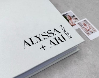 Puede incluir: Un álbum de fotos de boda de color verde claro con textura de lino con los nombres "ALYSSA + ARI" y la fecha "09.09.2023" impresos en negro. Dos fotos pequeñas están dentro del álbum. El álbum está sobre una superficie gris.