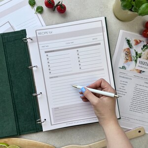 A recipe journal page with sections for recipe name, prep time, cook time, servings, ingredients, directions, and notes.  The page features a minimalist design with a light beige background and dark gray text.  A black pen rests on the page, suggesting the process of writing a recipe.  The page is part of a binder, with visible ring-bound edges. A4 binder size with 4 rings.