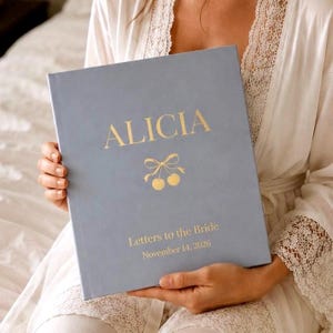 Puede incluir: Un libro de tapa dura azul claro con letras doradas y un gráfico de cerezas. La portada presenta el nombre ALICIA, el texto Letters to the Bride y la fecha November 14, 2026, centrados en un diseño limpio y sencillo en la parte frontal.