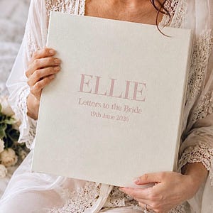 Puede incluir: Un libro de recuerdos cuadrado cubierto de tela color crema con el nombre ELLIE impreso en letras grandes de color rosa empolvado. Debajo del nombre, el texto dice Letters to the Bride y 19th June 2026 en un color de fuente a juego.