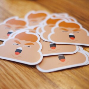 May include: A stack of brown and white poop emoji stickers with a red mouth and orange hair. The stickers are on a wooden surface.
