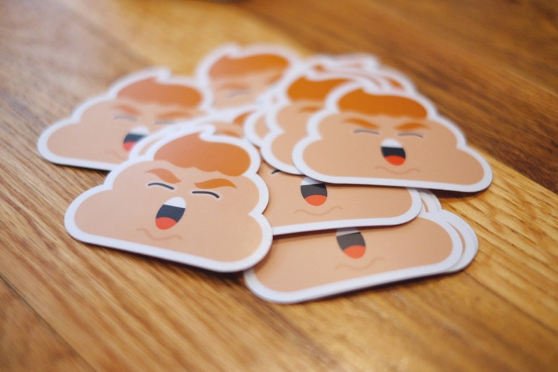 May include: A stack of brown and white poop emoji stickers with a red mouth and orange hair. The stickers are on a wooden surface.